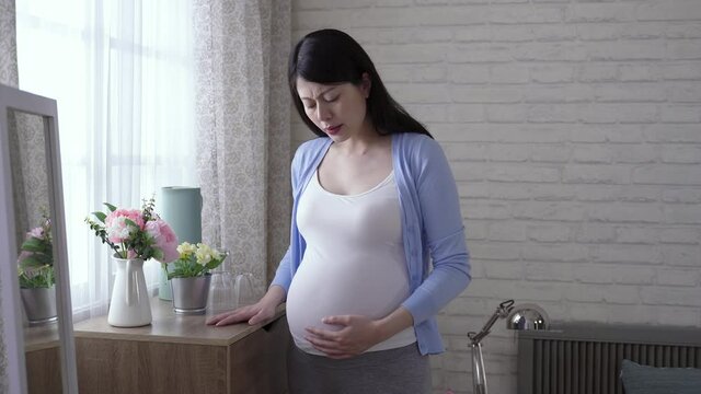pregnant woman walking to the home window is feeling a sudden contraction with her face contorted in a grimace of pain in the morning.