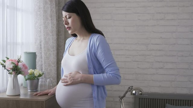 asian woman in the last trimester is feeling sudden pain in the abdomen in the morning by the window at home.
