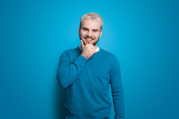 Obraz premium Man with blonde hair on blue wall is touching chin. Smiling man. Front view. Handsome man. Positive person.