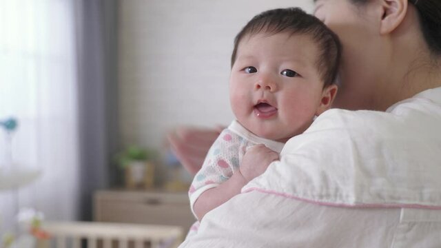 selective focus cute newborn infant is looking at camera innocently as her mommy is helping her burp at blurred background bright nursery room.