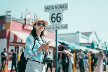 young elegant female tourist with sunglasses and hat holding mobile phone standing in amusement...