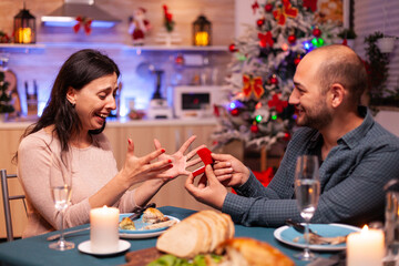 Happy man surpsing girlfriend with diamond luxury expensive engagement ring celebrating christmas holiday together sitting at dining table in xmas kitchen. Couple enjoying winter season