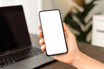 Close up of hand holding smartphone with white mock-up screen in home office..