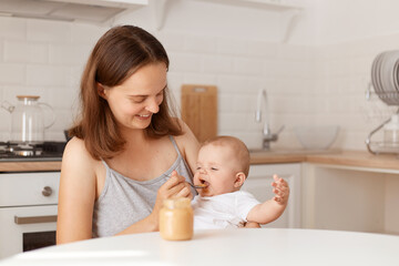 Happy dark haired woman feeding her little daughter with fruit or vegetable puree, sitting at table in kitchen, looking smiling at baby, expressing love and care, indoor shot.