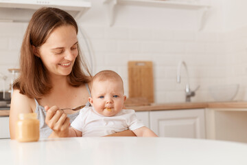 Smiling dark haired female feeds her little daughter with fruit or vegetable puree, sitting in light room with kitchen set on background, first feeding up.