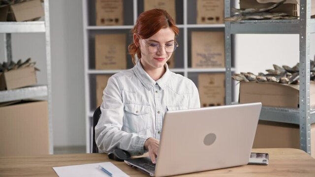 pretty young woman works in fish warehouse and takes orders online while sitting at table with laptop on background of shelves, smiling and looking at camera