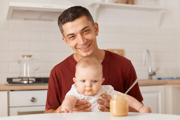 Obraz na płótnie Canvas Portrait of handsome father wearing burgundy t shirt with charming infant daughter, feeding up her toddler girl, looking smiling at camera, expressing happiness.