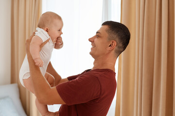 Side view of brunette man throwing up little daughter while standing in living room with window on background, happy parenthood, father spending time with child.