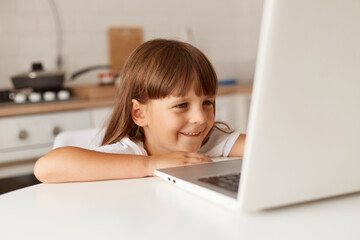 Smiling happy little preschooler girl with dark hair sitting in front of laptop computer in kitchen, looking at display, expressing positive emotions, watching cartoons.