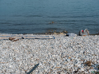 Beach cane mat, woman bag tote, white sandals on pebble seaside near water line surface front view