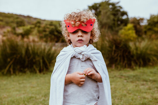 Cute blond boy with curly hair wearing mask and cape standing on meadow