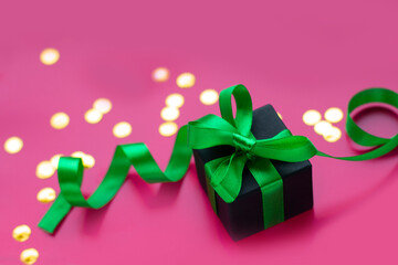 Gift box with green ribbon bow on light pink background with gold confetti. Festive decor background. Top view.