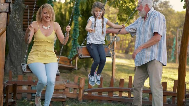 Slow motion shot of a smiling grandfather rocking his daughter and granddaughter on swings in the backyard. Three generations family spend time together swinging on the children’s playground