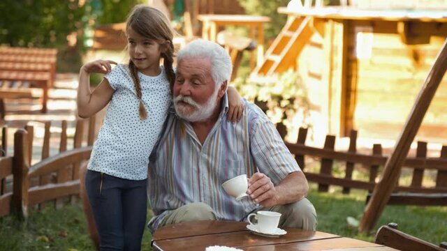 Sweet little girl hugs and kisses her grandfather in the yard while they spend time together. Elderly man drinks coffee in the yard and spends time with his granddaughter playing in the yard at home