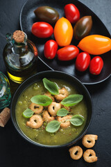 Bowl of vegetable soup topped with italian tarallini and fresh green basil, high angle view on a black stone background, vertical shot