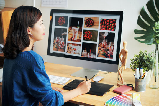 Close-up of busy young Asian designer sitting at wooden desk with color swatch and using digitizer while retouching photos