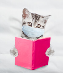 Kitten wearing medical protective mask lying under white warm blanket on a bed at home and reads a...
