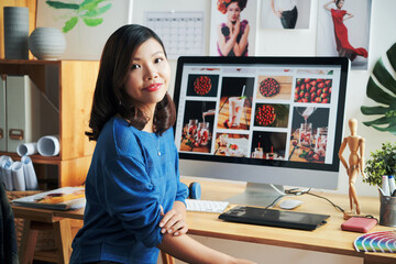 Fototapeta na wymiar Portrait of smiling young Asian stock photo designer in blue sweater sitting at modern computer in office