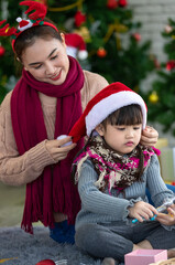 Attractive Asian mother and daughter wearing winter outfits and Santa's hat celebrating Christmas with joy and happiness with decorated Christmas tree background. Concept for family Christmas party