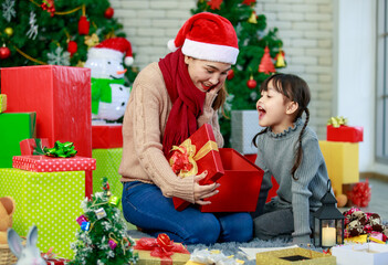 Obraz na płótnie Canvas Attractive Asian mother and daughter wearing winter outfits and Santa's hat unboxing gifts celebrating Christmas with joy. Decorated Christmas tree background. Concept for family Christmas party
