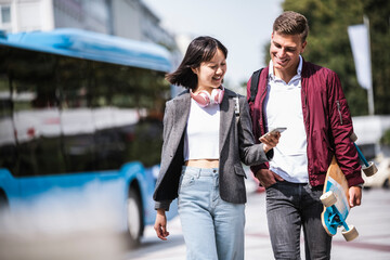 Smiling male and female friends sharing mobile phone while walking during sunny day