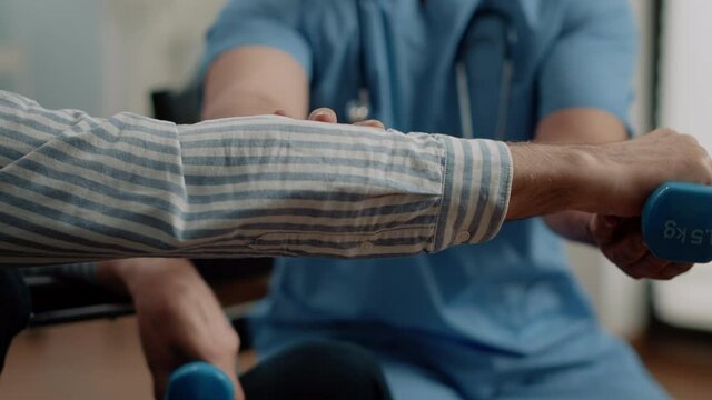 Close up of hand of patient holding dumbbells for physical activity and exercises while man nurse giving support with recovery. Disabled person doing mobility therapy in nursing home