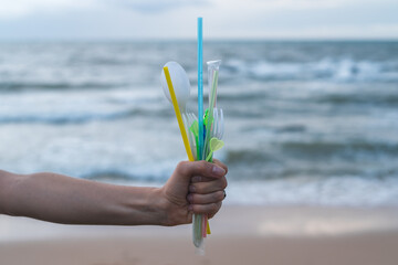 Fototapeta na wymiar One hand holding used plastic dishes and straws on the beach. Clean ocean beach on the background. Plastic pollution, Environmental Problem. 