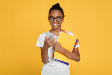 Smiling teenager african american girl pupil in glasses and casual holds books and reads message on...