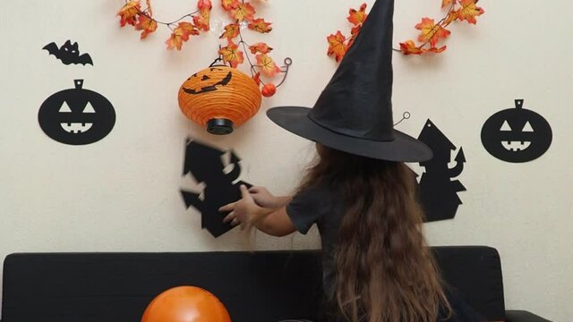 mom and daughter in fancy dress at home preparing for halloween. Girl in a witch hat. inflate balloons, prepare decorations. girl with devil horns carves bats and ghost, pumpkin