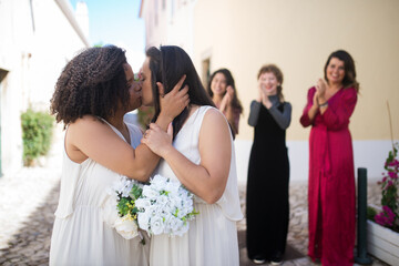 Portrait of cute kissing brides. Two young women hugging, kissing tenderly. Female guests laughing and applauding in background. Wedding, LGBT, celebration concept