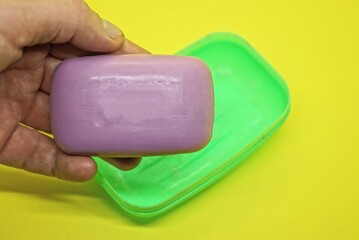hand holds one purple soap over a green plastic soap dish on a yellow table