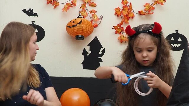 mom and daughter in fancy dress at home preparing for halloween. Girl in a witch hat. inflate balloons, prepare decorations. girl with devil horns carves bats and ghost, pumpkin