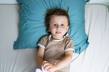 Naklejka premium Close up portrait of a child lying in a bed in the morning. Adorable toddler wearing pajamas waking up and playing, view from above. Easy awakening concept.