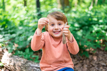 Little preschool boy sitting on a tree trunk in a forest and playing with wooden stick. Child spending time outdoor in summer. Toddler in a park. Ecological playground.