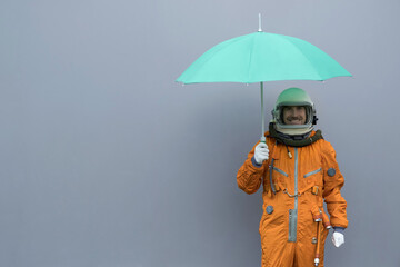 Happy astronaut wearing orange space suit and space helmet holding open green umbrella against gray...