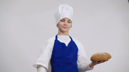 Proud little baker holding freshly-baked bread showing thumb up smiling looking at camera. Caucasian boy in apron and chef hat posing at white background with pastry. Cuisine and culinary profession