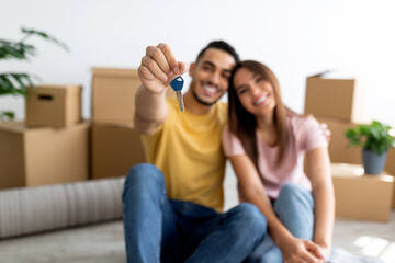 Positive young multiracial couple showing house key, sitting on floor of new home, selective focus