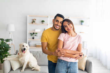 Portrait of happy multiracial couple hugging and smiling at camera, their adorable pet dog sitting on couch nearby