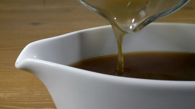 Closeup POV shot of hot gravy being poured from a glass measuring jug into a white serving boat.