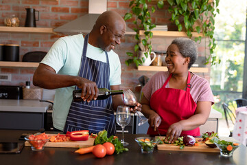 Happy african american senior couple cooking together in kitchen, drinking wine