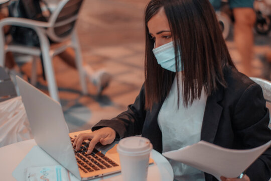 Profesora con mascarilla escribiendo en su laptop mientras corrige ex&aacute;menes en la cafeter&iacute;a y toma caf&eacute; por la ma&ntilde;ana 