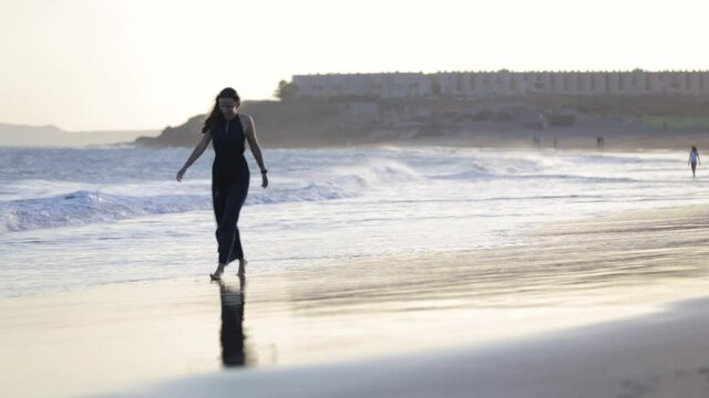Young woman in long evening dress walking along atlantic ocean beach at sunset.