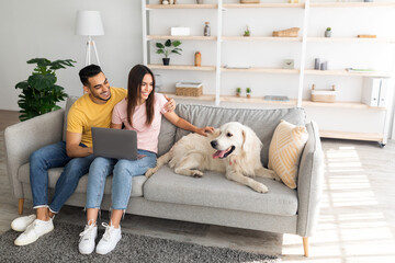 Cheerful young multiracial couple sitting on sofa with laptop, stroking their dog at home, copy space