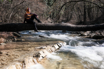 A girl sitting on the log lying above mountain forest river on sunny winter day. Krasnodar Krai, Caucasus, Russia.