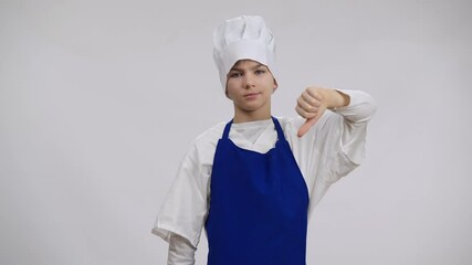 Caucasian boy in blue apron and white chef hat posing at background showing thumb down. Portrait of kid dissatisfied with culinary profession choice looking at camera shaking head no gesture