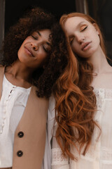 Fototapeta premium Beautiful African and European woman are standing next to each other with their heads touching outdoors. Eyes are closed on their dreamy faces, both are dressed in white clothes.