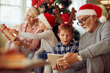 Senior man and his grandson use touchpad while his wife and other grandson are opening Christmas presents in the background at home. - obrazy, fototapety, plakaty