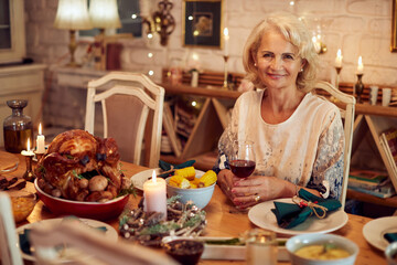 Happy senior woman enjoys in glass of wine at dining table on Thanksgiving.