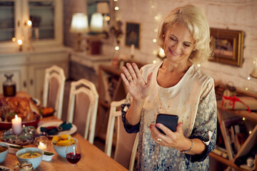 Happy mature woman uses smart phone and greets someone during video call on Thanksgiving.