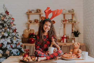 cute little brunette girl in checkered red pajamas with reindeer horns on her head is eating a Christmas cake and drinking tea from a white mug in a beautifully decorated kitchen. lifestyle, hygge